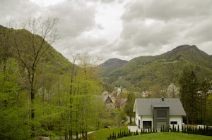 A modern house with a grey roof is situated in a lush green valley surrounded by dense trees and hills. In the background, taller mountains rise under a cloudy sky. A small path leads to the house, and a few more buildings with steep roofs can be seen in the distance.