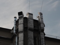 Several metallic ventilation ducts are attached to the exterior of a building, extending vertically. The sky in the background is overcast, and there are cables running across the scene.