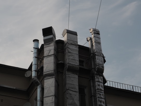 Several metallic ventilation ducts are attached to the exterior of a building, extending vertically. The sky in the background is overcast, and there are cables running across the scene.