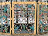Intricately designed wrought iron gate with vivid colors and ornate patterns, featuring a series of stylized animals, floral motifs, and scrollwork. The dominant colors are teal, yellow, and red, with the gate framed by gilded panels accentuated with detailed blossoms and curlicues.