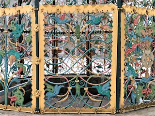 Intricately designed wrought iron gate with vivid colors and ornate patterns, featuring a series of stylized animals, floral motifs, and scrollwork. The dominant colors are teal, yellow, and red, with the gate framed by gilded panels accentuated with detailed blossoms and curlicues.