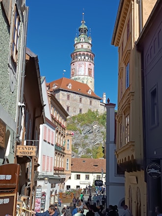 A picturesque street scene featuring a historic tower with ornate architectural details, flanked by colorful buildings. People stroll along the cobblestone path, and shop signs, including a candy shop, add to the charming atmosphere.