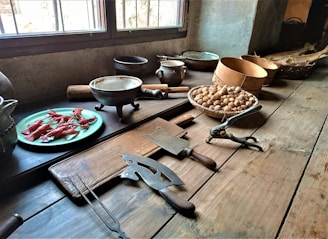 A rustic kitchen setting features a wooden table with various cooking tools and ingredients. There is a plate of red crustaceans, possibly crayfish, a selection of bowls and pots, a basket filled with walnuts, and several vintage kitchen tools including a cutting board, cleaver, and nutcracker.