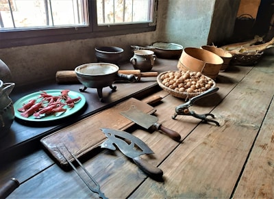 A rustic kitchen setting features a wooden table with various cooking tools and ingredients. There is a plate of red crustaceans, possibly crayfish, a selection of bowls and pots, a basket filled with walnuts, and several vintage kitchen tools including a cutting board, cleaver, and nutcracker.