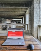 A modern laptop on a wooden table placed in an industrial-style hallway with stone walls and concrete beams. The laptop screen displays a webpage with a red header. A wireless mouse is positioned next to the laptop.