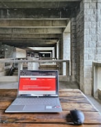 A modern laptop on a wooden table placed in an industrial-style hallway with stone walls and concrete beams. The laptop screen displays a webpage with a red header. A wireless mouse is positioned next to the laptop.