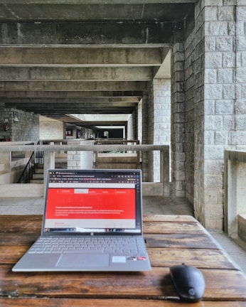 A modern laptop on a wooden table placed in an industrial-style hallway with stone walls and concrete beams. The laptop screen displays a webpage with a red header. A wireless mouse is positioned next to the laptop.