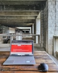 A modern laptop on a wooden table placed in an industrial-style hallway with stone walls and concrete beams. The laptop screen displays a webpage with a red header. A wireless mouse is positioned next to the laptop.