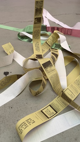 Long strips of paper featuring printed text and images are scattered on a concrete surface in a seemingly random manner. The papers come in various colors, including yellow, green, and white, and are curling and overlapping each other.