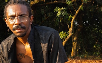 A person with braided hair and glasses is partially visible in a natural setting with trees and foliage in the background, under warm sunlight.