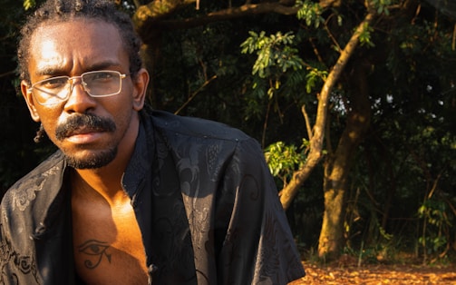 A person with braided hair and glasses is partially visible in a natural setting with trees and foliage in the background, under warm sunlight.