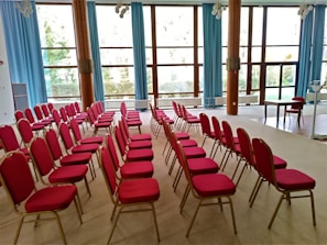 An empty conference room featuring rows of red chairs with gold frames arranged in a neatly organized pattern on a beige carpet. The room has large windows with blue curtains, allowing natural light to illuminate the space. A wooden table and chair are placed near the front of the room, suggesting a setup for presentations or speeches.