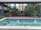 A tranquil outdoor space features a small rectangular fountain with clear blue water and cascading streams. The background includes a modern building with large glass windows surrounded by lush greenery, potted plants, and colorful flowers.