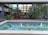 A tranquil outdoor space features a small rectangular fountain with clear blue water and cascading streams. The background includes a modern building with large glass windows surrounded by lush greenery, potted plants, and colorful flowers.