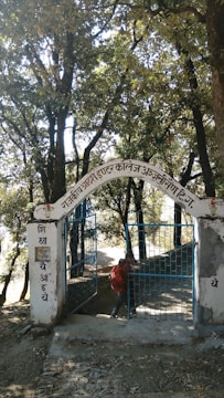 An iron gate with blue bars stands open, set within a white stone archway. The archway displays text in a local script. The background is filled with tall trees and dense foliage, casting shadows on the path leading through the gate. A person carrying a red backpack is visible, appearing to walk through the gateway.