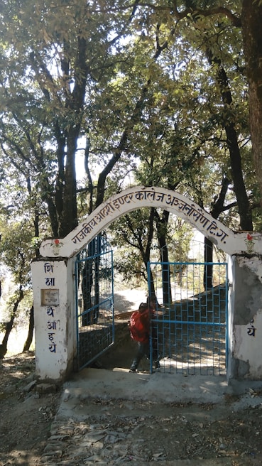 An iron gate with blue bars stands open, set within a white stone archway. The archway displays text in a local script. The background is filled with tall trees and dense foliage, casting shadows on the path leading through the gate. A person carrying a red backpack is visible, appearing to walk through the gateway.