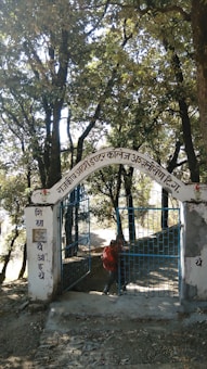 An iron gate with blue bars stands open, set within a white stone archway. The archway displays text in a local script. The background is filled with tall trees and dense foliage, casting shadows on the path leading through the gate. A person carrying a red backpack is visible, appearing to walk through the gateway.