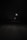 A dark road at night is illuminated by a series of distant streetlights. The road is mostly enveloped in shadows, creating a mysterious and somewhat eerie atmosphere. The faint outlines of trees and power lines are visible along the sides of the road.