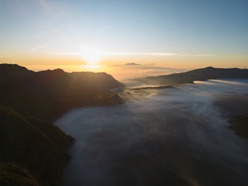 A breathtaking sunrise over a mountainous landscape with mist covering the valleys. The sun is at the horizon, casting a warm glow across the sky and creating long shadows over the terrain. The sky transitions from a deep blue to a soft orange at the horizon, adding to the serene atmosphere.