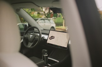 Interior view of a modern car, featuring a large touchscreen display mounted on the dashboard. The steering wheel is branded with a distinctive logo, and there's a clear view of another car through the window.