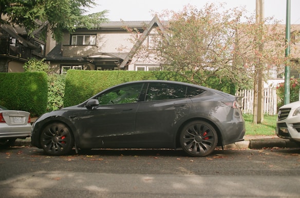 A sleek, dark-colored electric car is parked on a street flanked by lush greenery and trees. The vehicle is situated between two other cars. In the background, there is a residential house partially obscured by trimmed hedges. The setting has a peaceful suburban feel.
