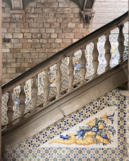 A stone staircase is adorned with intricate decorative tiles featuring floral patterns and vibrant colors. The tiles showcase a blend of blue, yellow, and white hues, set against the warm tones of the brick wall that forms the backdrop. The railing is comprised of ornate balusters, each carved with delicate detail.