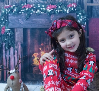 A little girl in a sparkly red dress with white fur trim, sitting by a fireplace with stockings hung above.