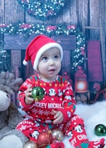 A baby wearing a red Santa hat and festive red pajamas with Mickey Mouse designs is sitting on a fluffy white surface. The baby is holding a green bauble and is surrounded by other colorful Christmas ornaments. In the background, there is a Christmas wreath, a wooden fireplace, and decorative garlands with white and red berries.