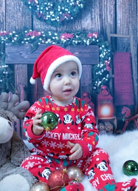 A baby wearing a red Santa hat and festive red pajamas with Mickey Mouse designs is sitting on a fluffy white surface. The baby is holding a green bauble and is surrounded by other colorful Christmas ornaments. In the background, there is a Christmas wreath, a wooden fireplace, and decorative garlands with white and red berries.