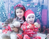 Two children dressed in festive outfits are positioned in front of a holiday-themed backdrop. The older child is holding a plush reindeer toy, while the younger child wears a Santa hat and holds Christmas ornaments. In the background, there are wreaths and decorative elements suggesting a cozy, wintry setting.