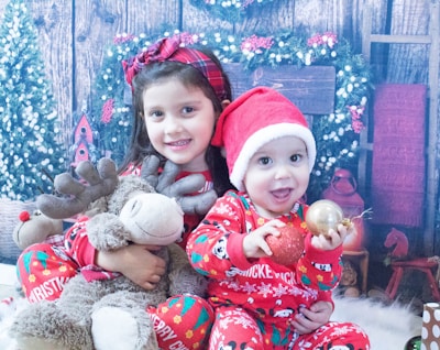 Two children dressed in festive outfits are positioned in front of a holiday-themed backdrop. The older child is holding a plush reindeer toy, while the younger child wears a Santa hat and holds Christmas ornaments. In the background, there are wreaths and decorative elements suggesting a cozy, wintry setting.