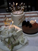 Three elegantly decorated cakes are placed on a table. The first cake has intricate golden decorations resembling a crown with pearl accents. The second cake is adorned with large white flower decorations and gold beads. The third cake appears to be chocolate with a 'Happy Birthday Wajira' topper and a candle. The tablecloth is patterned with light and dark brown stripes.