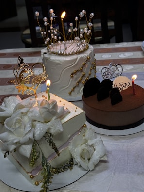 Three elegantly decorated cakes are placed on a table. The first cake has intricate golden decorations resembling a crown with pearl accents. The second cake is adorned with large white flower decorations and gold beads. The third cake appears to be chocolate with a 'Happy Birthday Wajira' topper and a candle. The tablecloth is patterned with light and dark brown stripes.