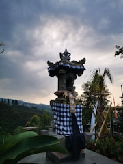 A traditional Balinese shrine with an intricately carved roof stands decorated with a checkered cloth and ornamental details. The structure is set against a backdrop of lush greenery and hills under a cloudy sky.