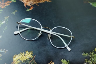 A pair of round eyeglasses with clear lenses and white frames rests on a dark surface. The background features decorative patterns of leaves and pine cones, displaying hints of green, brown, and orange colors.