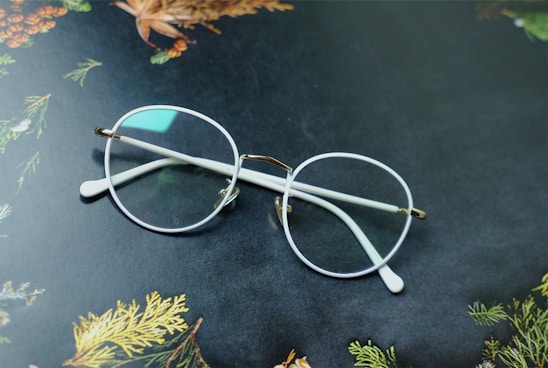 A pair of round eyeglasses with clear lenses and white frames rests on a dark surface. The background features decorative patterns of leaves and pine cones, displaying hints of green, brown, and orange colors.