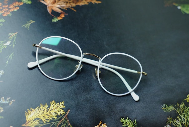 A pair of round eyeglasses with clear lenses and white frames rests on a dark surface. The background features decorative patterns of leaves and pine cones, displaying hints of green, brown, and orange colors.