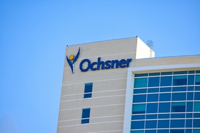A modern building with a beige brick facade and large blue-tinted windows. The building displays a prominent logo and name in blue lettering on one side.