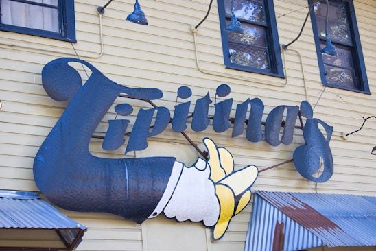 A whimsical sign features the word 'Tipitina's' prominently in stylized, metallic text attached to a yellow wooden building with horizontal siding. The sign includes a whimsical depiction of a hand tossing bananas. Blue industrial-style light fixtures and windows with reflective glass are seen above the sign.