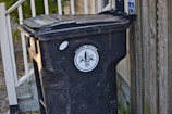 A black trash bin with a lid is positioned next to a wooden fence and a railing with leaves scattered on the ground. The bin is labeled 'City of New Orleans', featuring a fleur-de-lis design.