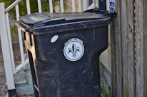 A black trash bin with a lid is positioned next to a wooden fence and a railing with leaves scattered on the ground. The bin is labeled 'City of New Orleans', featuring a fleur-de-lis design.