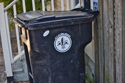 A black trash bin with a lid is positioned next to a wooden fence and a railing with leaves scattered on the ground. The bin is labeled 'City of New Orleans', featuring a fleur-de-lis design.