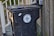 A black trash bin with a lid is positioned next to a wooden fence and a railing with leaves scattered on the ground. The bin is labeled 'City of New Orleans', featuring a fleur-de-lis design.
