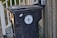 A black trash bin with a lid is positioned next to a wooden fence and a railing with leaves scattered on the ground. The bin is labeled 'City of New Orleans', featuring a fleur-de-lis design.