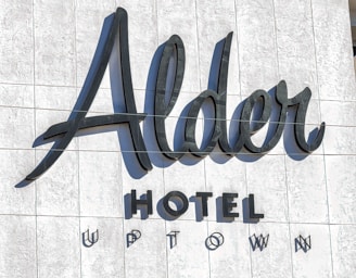 Large, stylish lettering spells out 'Alder Hotel Uptown' on a textured, light-colored wall. The design features a mix of elegant cursive and bold uppercase fonts, creating a modern and sophisticated look.