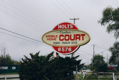 An outdoor sign for a mobile home sales and travel trailer site, featuring the text 'Holts Mobile Home Court' in bold red letters. The sign has a retro design, surrounded by trees and utility poles, under an overcast sky.
