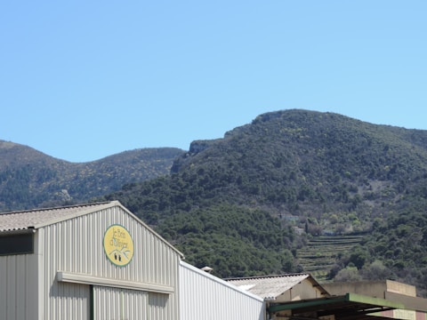 A large industrial building sits in front of a backdrop of rolling green hills under a clear blue sky. The building features a sign with the name 'Le Brin d'Olivier'. The mountainous terrain is covered in trees and vegetation, creating a serene natural setting.