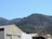 A large industrial building sits in front of a backdrop of rolling green hills under a clear blue sky. The building features a sign with the name 'Le Brin d'Olivier'. The mountainous terrain is covered in trees and vegetation, creating a serene natural setting.