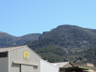 A large industrial building sits in front of a backdrop of rolling green hills under a clear blue sky. The building features a sign with the name 'Le Brin d'Olivier'. The mountainous terrain is covered in trees and vegetation, creating a serene natural setting.