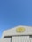A building with corrugated metal siding and a logo displaying 'Le Brin d'Olivier' under a clear blue sky. The logo has an image of olives and an olive branch.